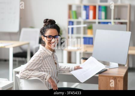 Smiling Young Hispanic Woman Working At Office Desk With Laptop And Documents, Business Concept Stock Photo