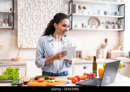 Happy Hispanic Woman Writing Shopping List While Cooking In Modern Kitchen, Healthy Lifestyle Concept Stock Photo