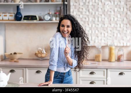 Smiling Young Woman With Curly Hair Holding Whisk In Modern Kitchen, Cooking Concept Stock Photo