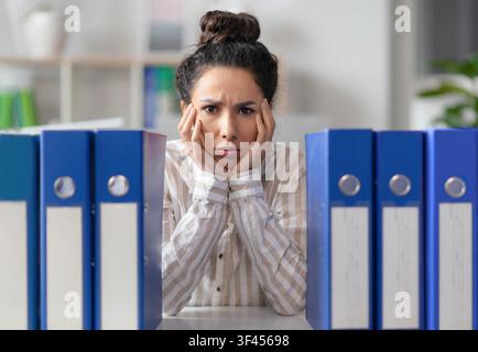 Worried Hispanic Woman Sitting At Desk With Folders, Facing Overwhelming Workload, Business Concept Stock Photo