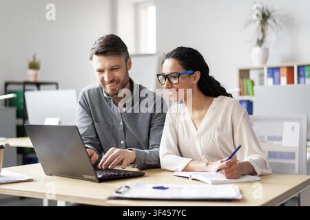 Smiling Black Woman Taking Notes While Collaborating With Man On Laptop In Modern Office Stock Photo