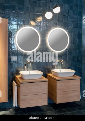 Contemporary bathroom showcasing two sinks, wooden vanities, and backlit mirrors against blue tiled wall Stock Photo