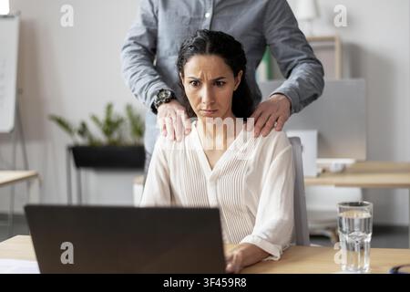 Concerned Hispanic Woman Working On Laptop With Overhead Hand Touch, Office Setting Stock Photo