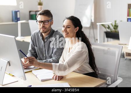 Smiling Hispanic Woman Assisting Caucasian Male Colleague Using Computer At Office, Business Concept Stock Photo