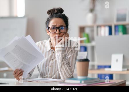 Worried Young Hispanic Woman Reading Documents At Desk In Office, Business Concept Stock Photo