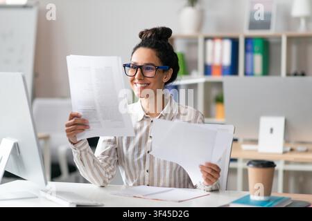 Smiling Hispanic Female Professional Reviewing Documents At Office Desk, Business Concept Stock Photo