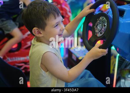 Boy playing arcade racing game in entertainment center. Child smiling while driving virtual car in amusement arcade. Excited kid having fun with video Stock Photo