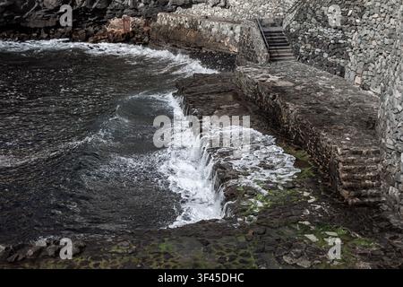 Charco Azul coastal stone terraces in La Palma feature rugged basalt steps leading down to ocean. Ocean waves crash violently against dark volcanic Stock Photo