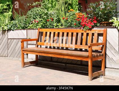 Wooden park bench garden photo. Empty varnished timber seat stands on brick pavement in front of blooming flowerbed with red orange blossoms Stock Photo