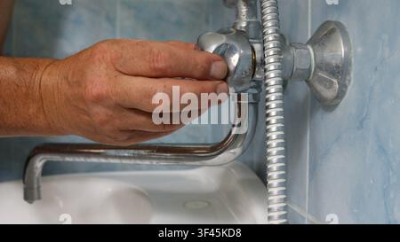 A man's hand adjusts or repairs a chrome faucet valve in a bathroom. The long spout and the edge of a white sink are visible below, and a shower hose Stock Photo