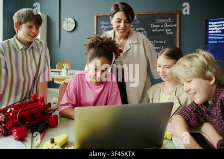 Female teacher guiding diverse school kids using laptop during coding and robotics lesson, supporting STEM teamwork and digital learning. Ideal for education technology promotion Stock Photo