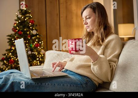 Young woman relaxes on a sofa using a laptop while holding a mug in a cozy living room with a decorated Christmas tree. Remote work, online shopping a Stock Photo