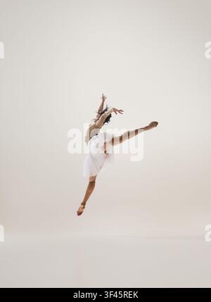 Young brunette ballerina performing split leap in white tutu in minimal studio Stock Photo