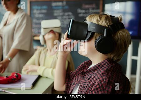 Caucasian schoolchildren wearing virtual reality headsets and exploring immersive lesson with teacher supervision in classroom. Ideal for education technology, STEM learning, digital training Stock Photo