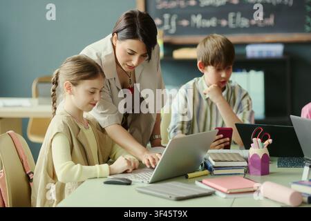 Caucasian female teacher guiding schoolchildren using laptop and smartphone during classroom lesson, supporting digital learning and teamwork. For education technology promotion and school marketing Stock Photo