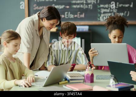 Caucasian female teacher guiding diverse children using laptops and tablet in classroom, supporting digital learning and coding education. Ideal for edtech, STEM, school marketing Stock Photo