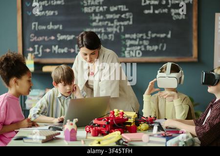 Teacher guiding diverse children using laptop and VR headsets during coding and robotics lesson in classroom. Ideal for STEM education, digital learning, innovation campaigns Stock Photo