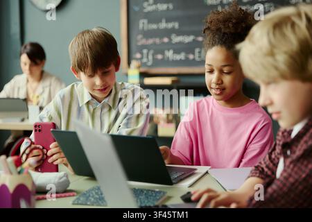 Multiethnic schoolchildren collaborating on laptops and smartphone in classroom, learning digital skills and teamwork. Ideal for education technology, coding class, STEM promotion Stock Photo