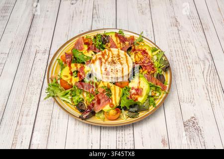 Grilled goat cheese salad with avocado, cured ham, cherry tomatoes, olives, and mixed greens on striped plate Stock Photo