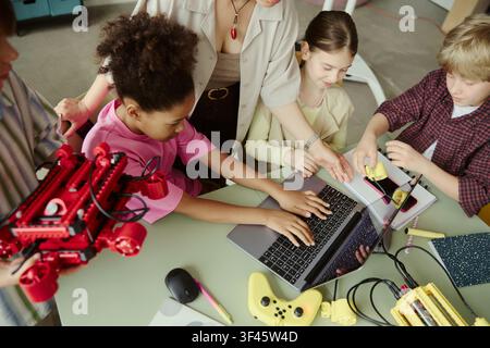 High angle view of diverse kids learning robotics, coding, collaborating with teacher around laptop and devices. Ideal for STEM education, digital skills, teamwork marketing Stock Photo