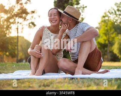 A smiling woman laughs as her partner lovingly kisses her cheek while they relax together in a peaceful park. Stock Photo