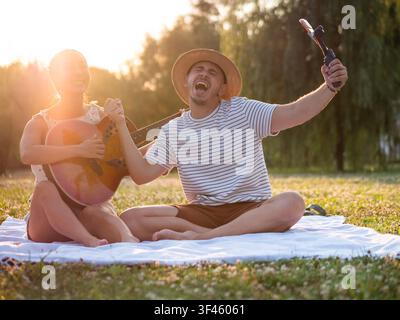 A smiling couple creates lasting memories during a relaxing picnic as they record a selfie video with a smartphone mounted on a handheld gimbal. Stock Photo
