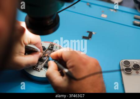 Technician repairs smartphone circuit board under magnifying lamp on blue work surface with tools and components visible in the background Stock Photo