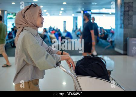 Side view of young Muslim female traveler pushing luggage trolley inside airport terminal. Stock Photo