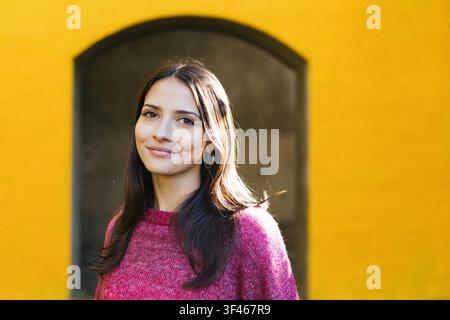 Young woman framed by arch smiling in front of a yellow wall Stock Photo