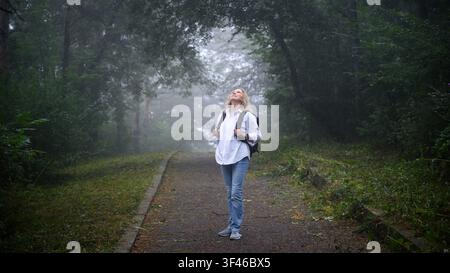 A blonde woman stands on an asphalt path in a dense, foggy forest, looking up at the trees. She is wearing a white button-down shirt, blue jeans, gray Stock Photo