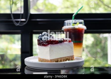 Slice of blueberry cheesecake with berry compote topping on white plate, iced honey lemon americano blurred in background by window Stock Photo