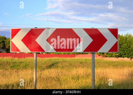 Horizontal chevron road sign with red and white arrows indicating sharp turn or detour, installed on metal poles near green trees and field under brig Stock Photo