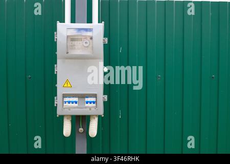 Outdoor electricity switchboard. Gray plastic switchboard with circuit breakers is mounted on a concrete pole against a green metal fence. Text space Stock Photo