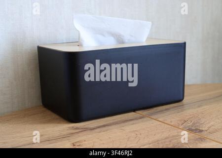 Black, minimalist paper napkin container sits on a table. White paper napkin protrudes from the black tissue box. Modern hygiene concept in a home int Stock Photo