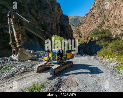 Heavy-duty yellow and grey excavator operates on gravel road within rocky mountain gorge. Steep cliffs and sparse vegetation frame the rugged scene un Stock Photo