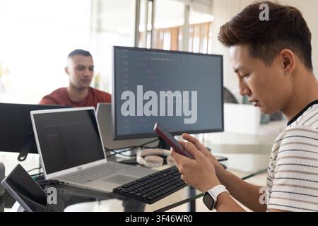 Diverse male coworkers using smartphone and coding on laptop with external monitor showing code Stock Photo