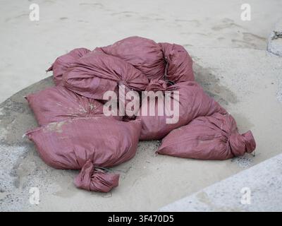 A pile of stacked purple-red woven plastic sacks filled with sand, tied at the ends and placed on a concrete surface near sand. Stock Photo