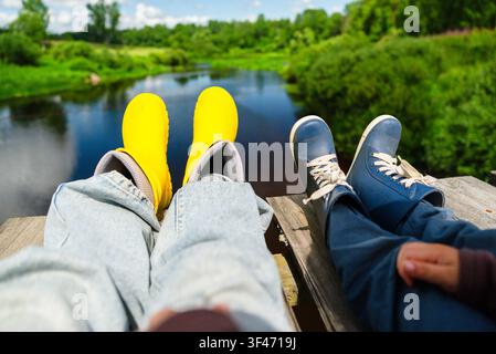 A mother and son sit on a wooden platform, dangling boots over the edge while enjoying a peaceful river view. This relaxed family moment captures serene happiness by the water. Stock Photo