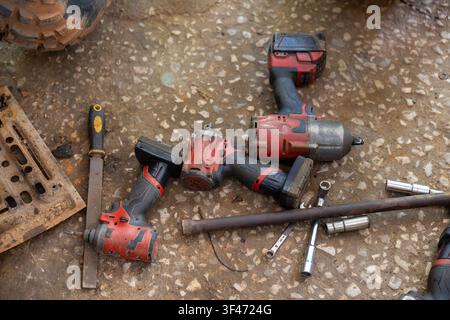 A worn red cordless impact wrench and drill on dusty workshop floor with sockets and metal bar, showing rugged repair scene and gritty texture Stock Photo