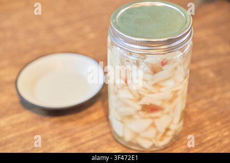 Pickled white cabbage and radish in a glass preservation jar beside an empty small dish on a wooden surface. Stock Photo