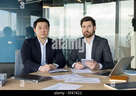 Two focused business partners collaborating at a desk, reviewing documents and using digital devices during a working session in a bright, contemporary workspace Stock Photo