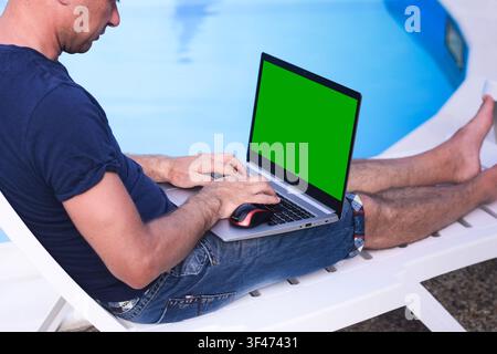 Laptop with male working outdoors on beach chair. laptop with green screen used for remote job and travel lifestyle, laptop showing chroma display for Stock Photo