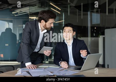 Business professionals collaborating in a meeting, reviewing financial charts and reports on a laptop and tablet, actively discussing strategy and project planning in a modern office environment Stock Photo