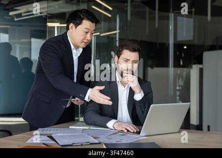 Diverse business partners discussing new strategy while working together at a desk, reviewing data and planning a project on a laptop in a contemporary workspace Stock Photo