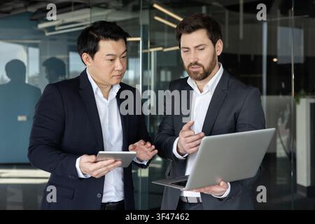 Two diverse businessmen standing in a modern office, collaborating on a silver laptop and a digital tablet, discussing strategies and innovative ideas for project success Stock Photo