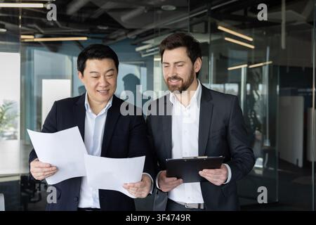 Two smiling businessmen, one asian and one caucasian, collaborating on a project, discussing financial reports while reviewing paperwork in a contemporary corporate environment Stock Photo