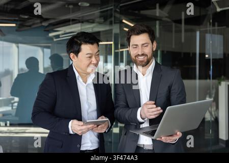 Two diverse businessmen standing in a modern office, smiling and actively discussing work, engaging with a laptop and a digital tablet, showcasing teamwork and professional collaboration Stock Photo