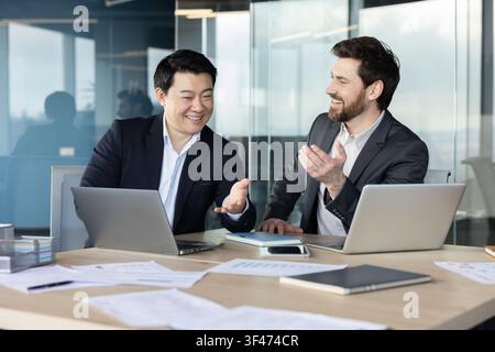 Two diverse businessmen collaborating and smiling during an office meeting, discussing projects while sharing ideas and using laptops at a conference table with documents Stock Photo