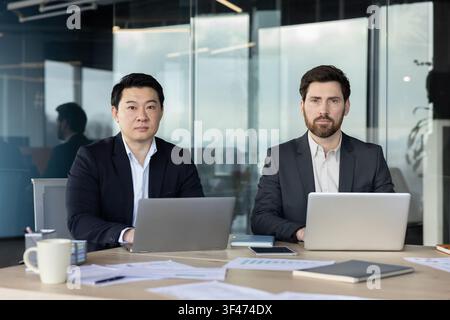 Two confident businessmen, one asian and one caucasian, sitting at a desk with laptops and paperwork, collaborating and working together in a contemporary corporate environment Stock Photo