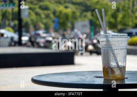 An almost empty disposable plastic cup with two straws sits on a round outdoor table, featuring a beautiful bokeh background of a sunny urban street w Stock Photo